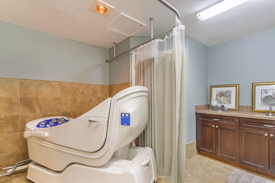 A bathroom with a large white assisted bathing tub featuring control buttons, beige tiled walls and floor, a shower curtain on a curved rod, and a wooden cabinet with a granite countertop holding framed pictures and a small mirror.