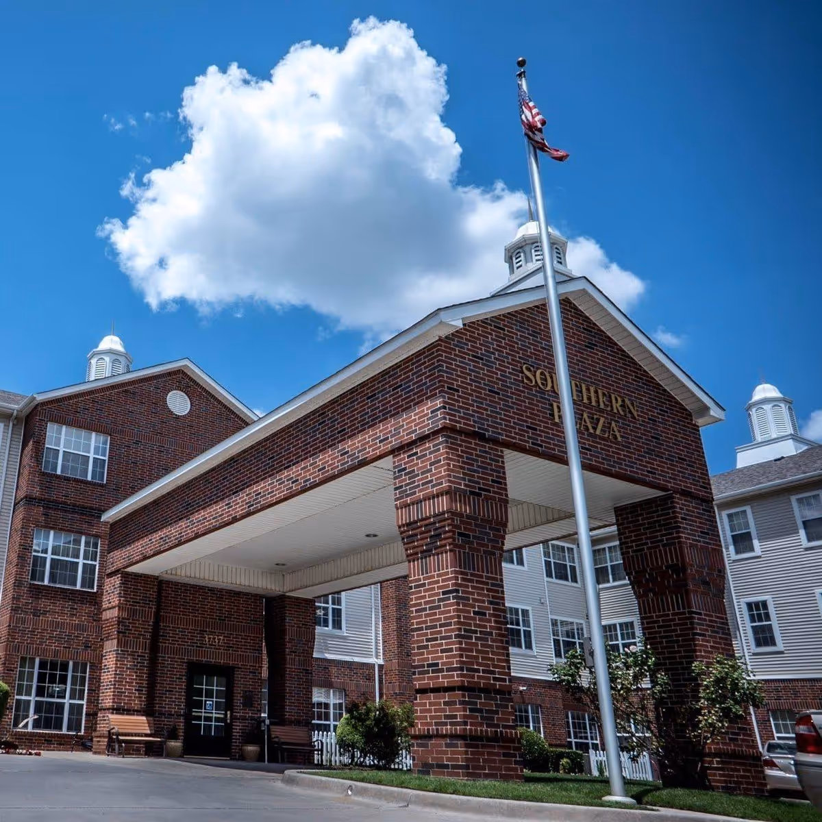 Exterior view of Southern Plaza Retirement Community building with a brick entrance canopy, an American flag on a flagpole, and a bright blue sky with a large white cloud.