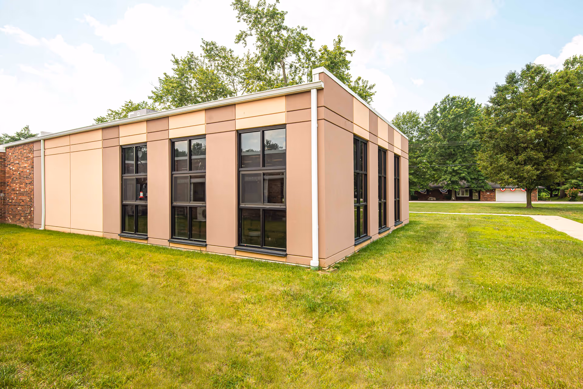 Exterior view of a single-story building with large windows and beige and brown paneling, surrounded by green grass and trees under a partly cloudy sky.