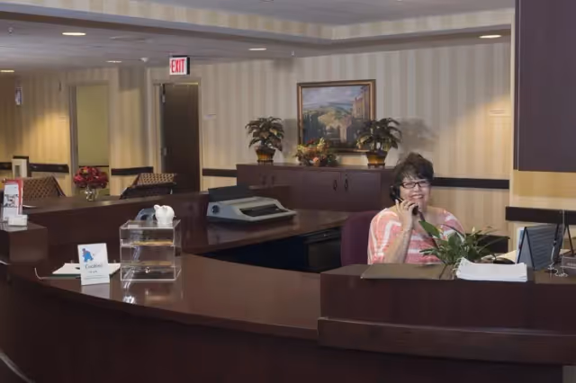 A woman sitting behind a dark wood reception desk in an assisted living facility, talking on the phone. The desk has a computer monitor, a plant, a clear container, and some papers. The background shows a hallway with an exit sign, framed artwork, and potted plants on a cabinet.