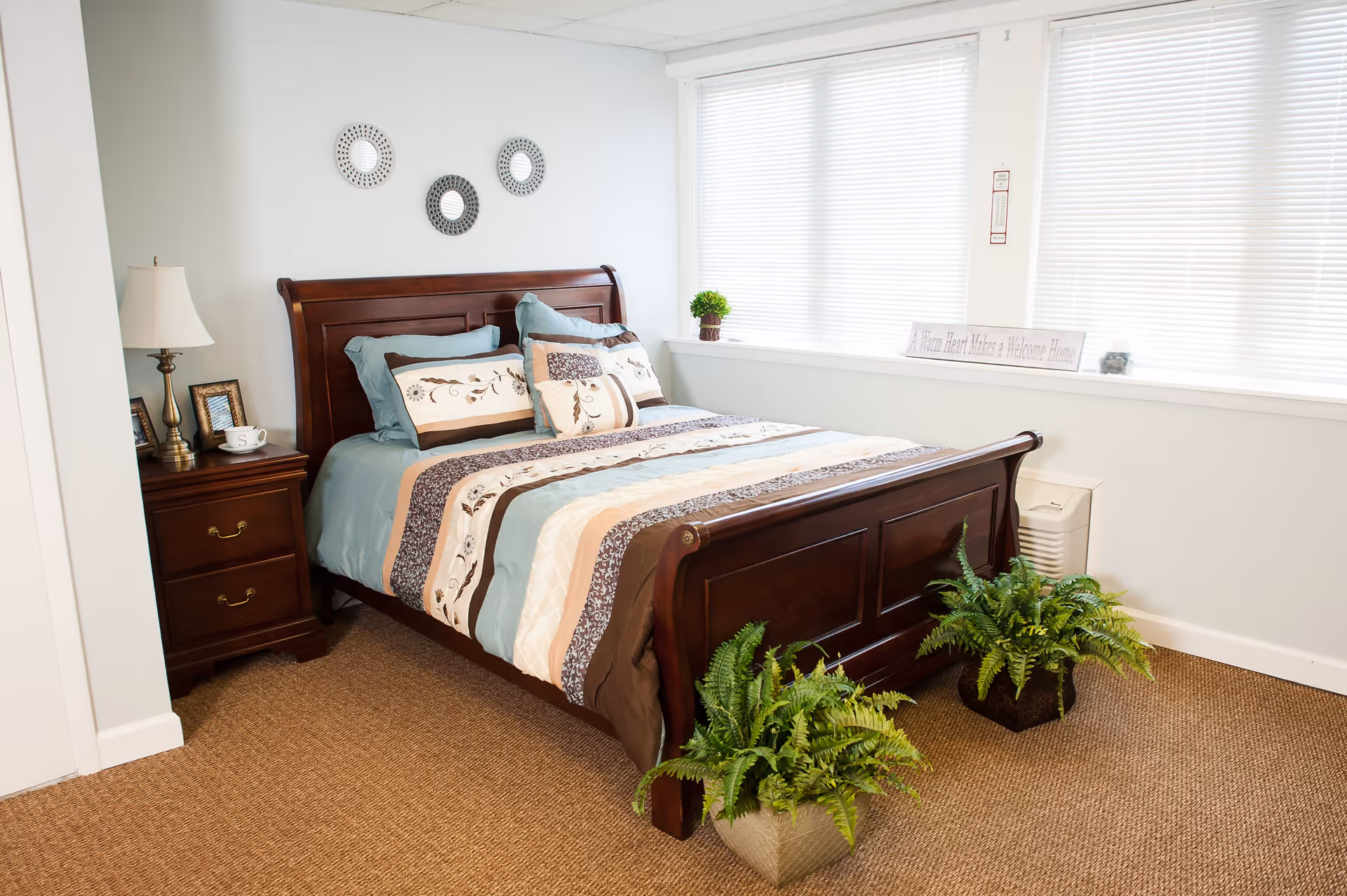 A neatly made bed with a wooden frame and a striped comforter in shades of blue, brown, and cream. There are multiple pillows arranged on the bed. On the left side, there is a wooden nightstand with a lamp, picture frames, and a cup. Two green potted plants are placed on the floor near the foot of the bed. The room has a large window with white blinds and a sign on the windowsill that reads 'A Warm Heart Makes a Welcome Home'. The walls are painted white and the floor is carpeted in a light brown color.
