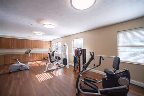 A well-lit exercise room with wooden flooring featuring various fitness equipment including a stationary bike, a multi-function weight machine, and an elliptical trainer. The room has two windows with blinds allowing natural light to enter, and wooden cabinets along one wall for storage.