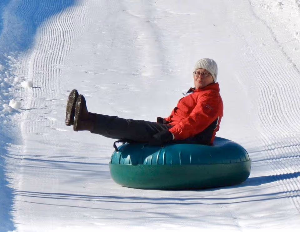 A person wearing a red jacket, black pants, boots, gloves, and a white knit hat is sitting on a green snow tube sliding down a groomed snowy hill.