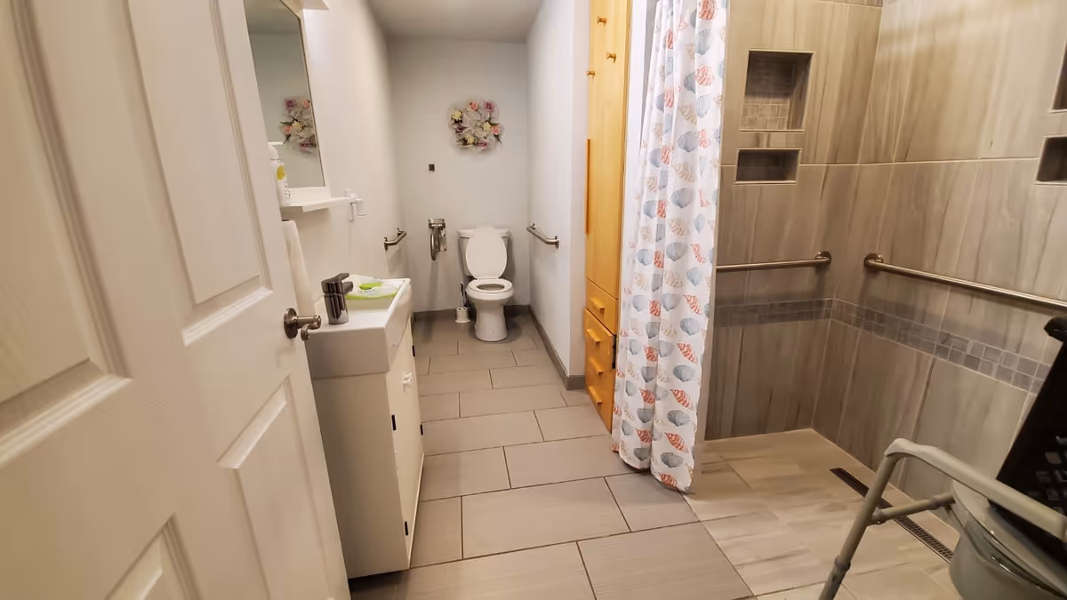 A clean and accessible bathroom featuring a toilet with grab bars on both sides, a small sink with a mirror above it, a wooden cabinet, and a walk-in shower with a patterned shower curtain and grab bars inside. The floor is tiled and the walls are light-colored.