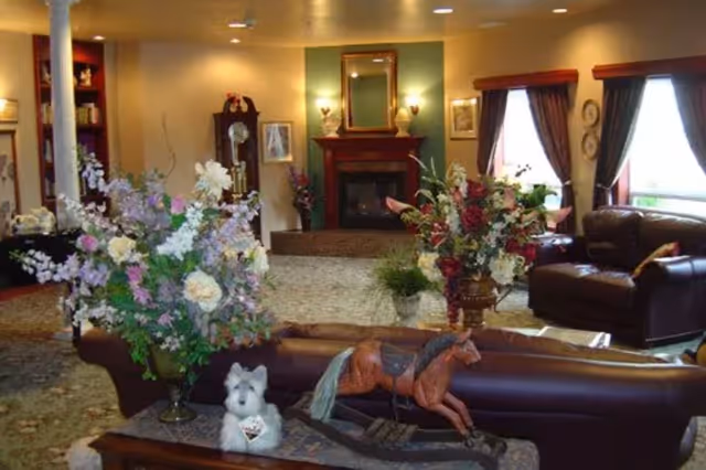 A cozy living room with a fireplace centered on the far wall, flanked by two lamps and framed pictures. The room features large windows with dark curtains, leather armchairs, and a sofa. In the foreground, there are two large floral arrangements and a wooden rocking horse decoration on a table, along with a small white stuffed dog.