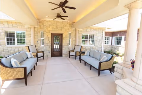 Covered outdoor seating area with two ceiling fans, two sofas, and four chairs arranged around a central door with stone walls and large windows on either side.