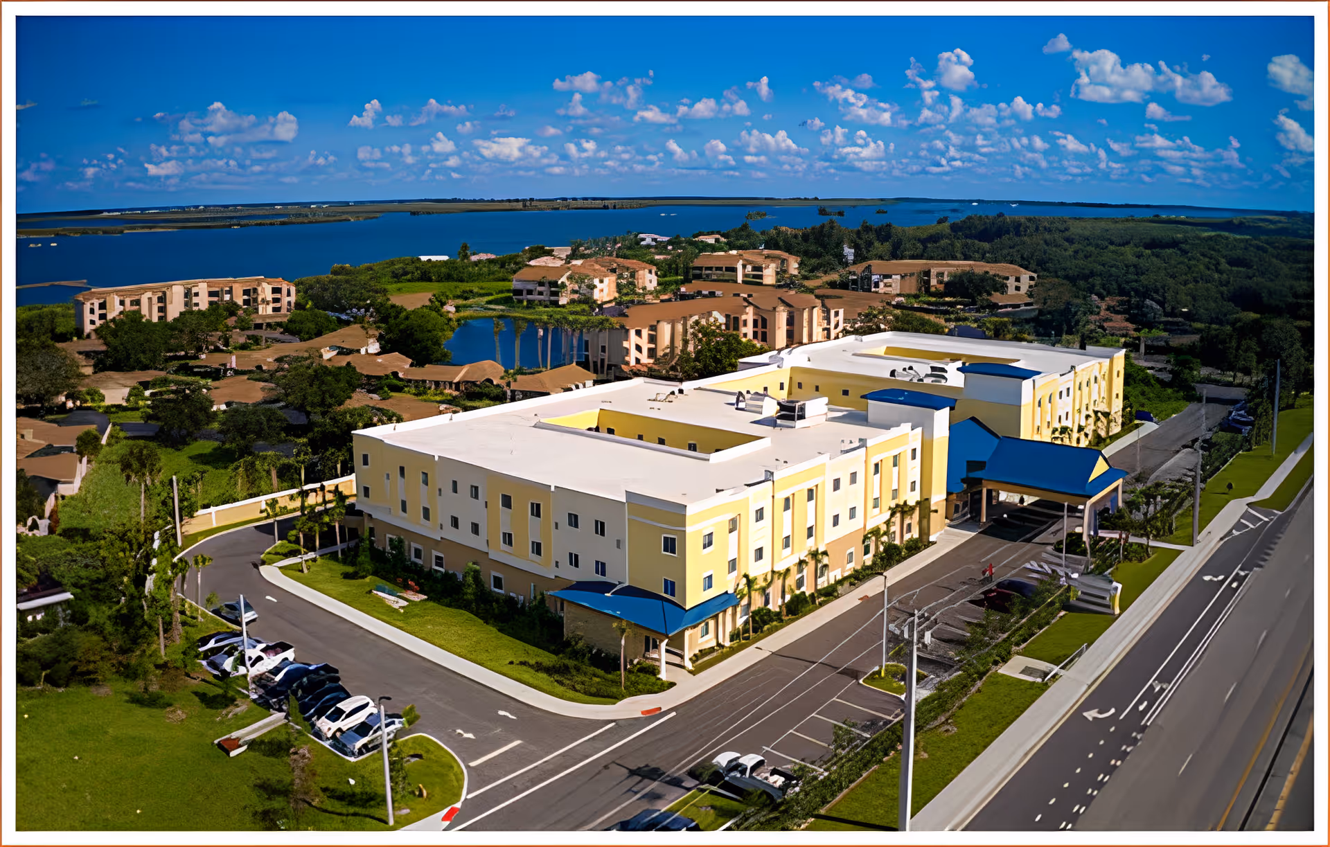 Aerial view of The Promenade senior living facility showing a large, three-story yellow and white building with blue awnings. The building is surrounded by parking lots, roads, green lawns, and trees. In the background, there are several other residential buildings, a pond, and a large body of water under a blue sky with scattered clouds.