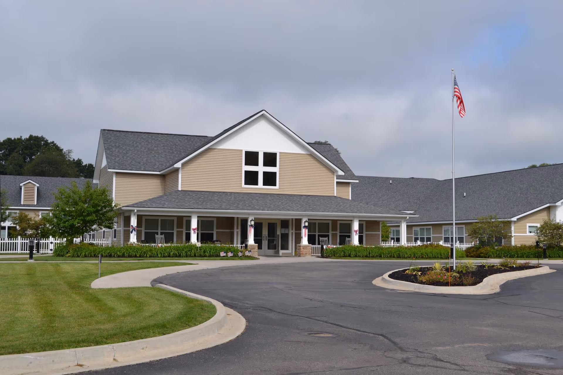 Front exterior view of Medilodge of Montrose, a two-story building with beige siding and a gray roof. The entrance has a covered porch with white columns and rocking chairs. There is a circular driveway with a landscaped island featuring an American flag on a flagpole. The surrounding area has green lawns and some trees under a cloudy sky.