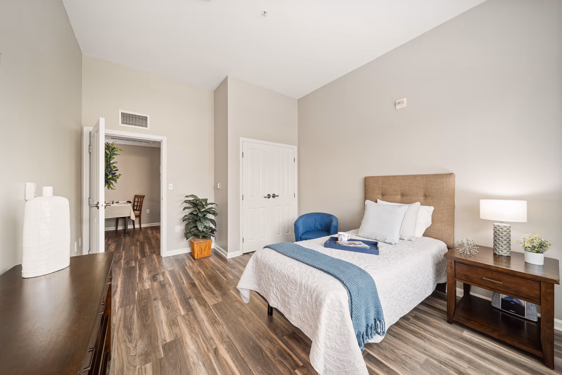 A bright bedroom with a single bed covered in white bedding and a blue throw blanket. There is a blue armchair next to the bed, and a wooden nightstand with a lamp, decorative items, and a small plant. The room has wood flooring, beige walls, a double-door closet, and an open door leading to another room with a dining table and chairs. A large white vase sits on a wooden dresser on the left side of the room.