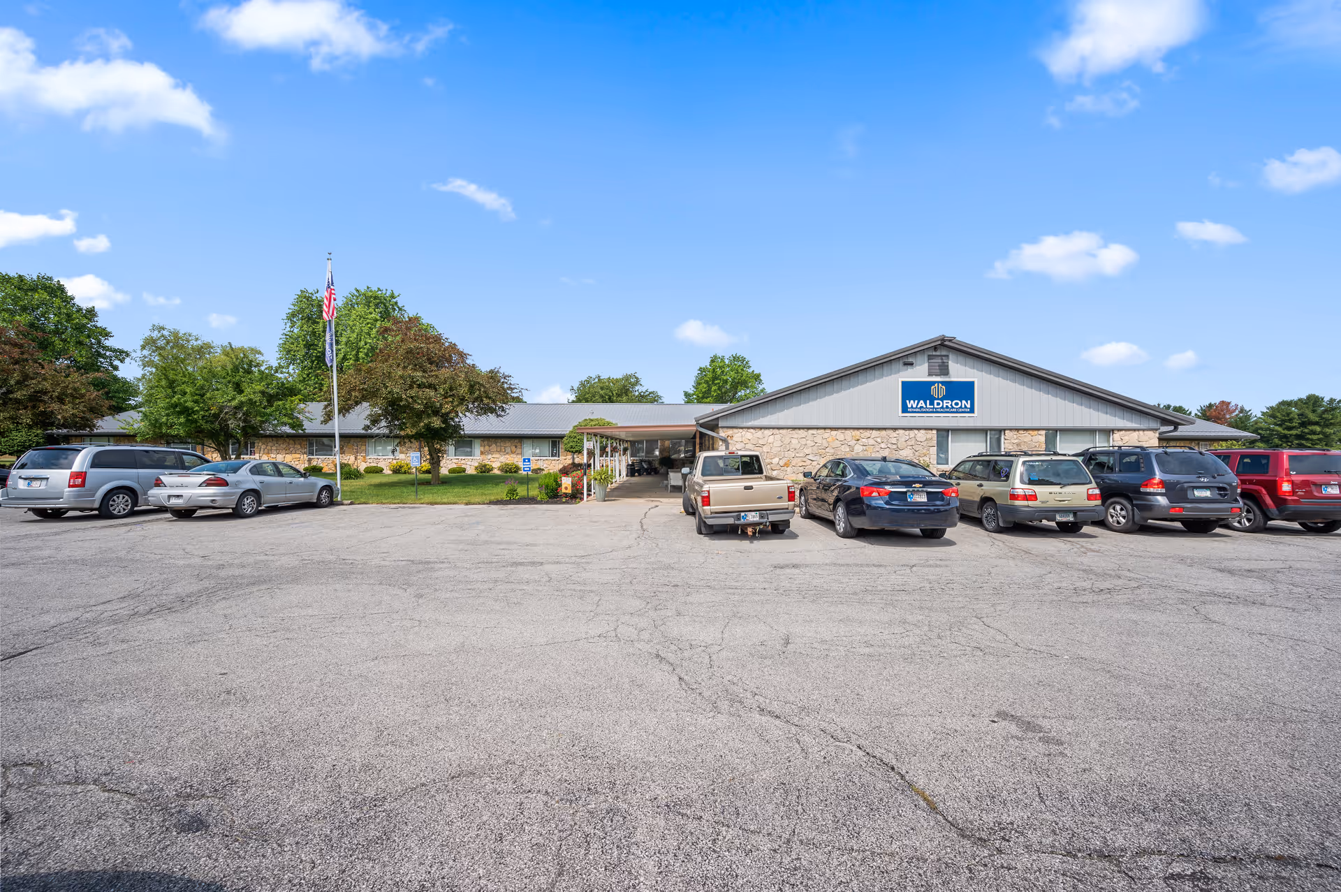 Exterior view of Waldron Rehabilitation and Healthcare Center building with a parking lot in front. Several cars are parked, and there are trees and a flagpole with the American flag near the entrance. The sky is clear with a few clouds.