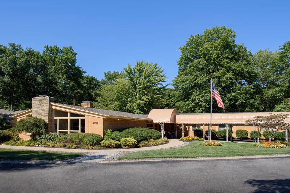 Single-story brick senior living facility with landscaped grounds, a covered entrance and an American flag on a flagpole.