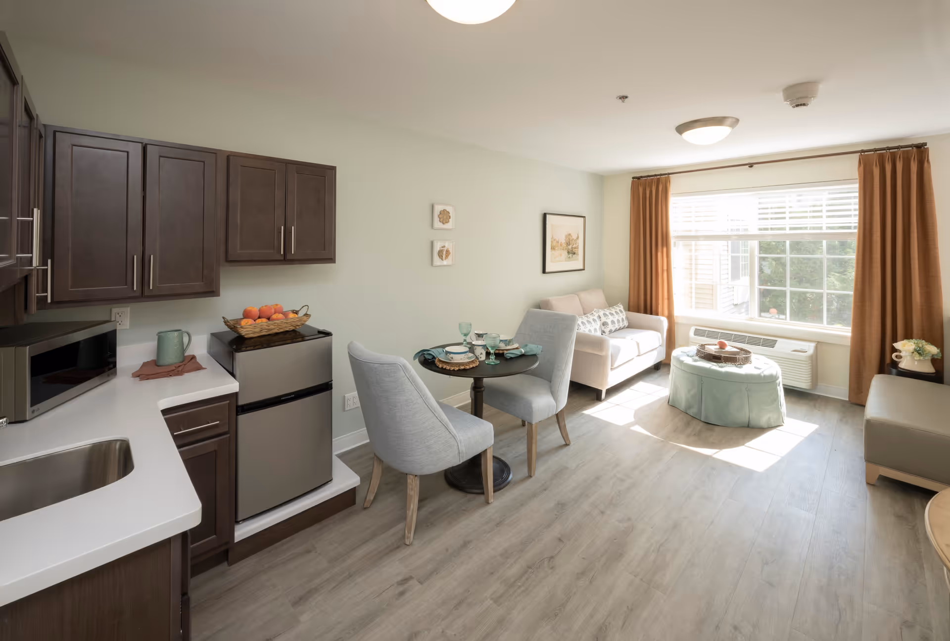 A bright and cozy living space in a senior living facility featuring a small kitchen area with dark wood cabinets, a microwave, and a mini refrigerator. Adjacent to the kitchen is a small round dining table set with two gray upholstered chairs and tableware. The living area includes a beige sofa, a round ottoman with a tray on top, and a large window with brown curtains allowing natural light to fill the room. The floor is covered with light wood laminate.