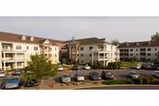 Exterior view of a multi-story senior living building with balconies, landscaped grounds, and a parking lot with parked cars.