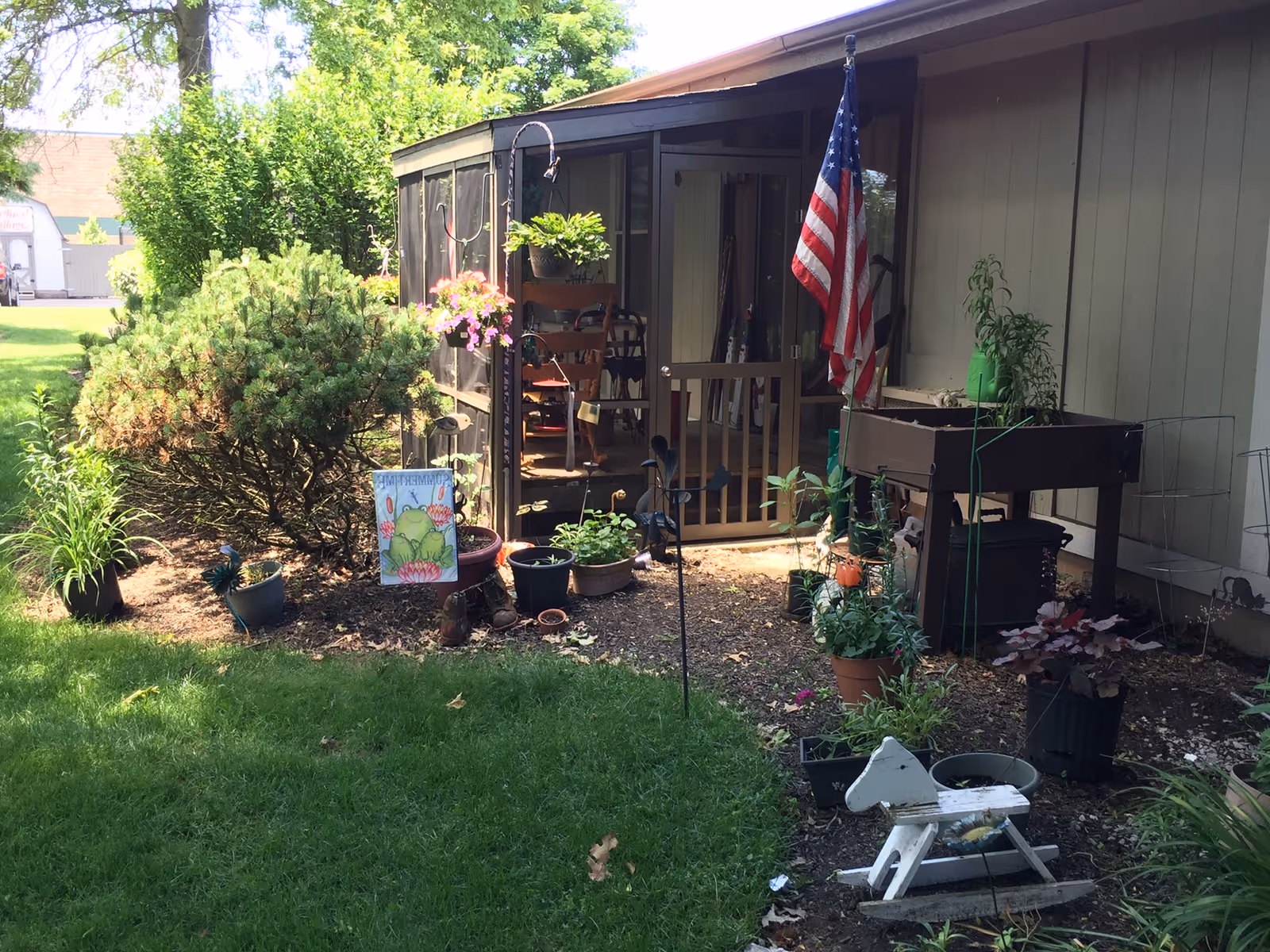 A garden area outside a building with various potted plants, a small bush, a wooden rocking horse, and an American flag mounted near a screened porch entrance. The porch has a wooden chair and some hanging plants, with green grass and trees visible in the background.