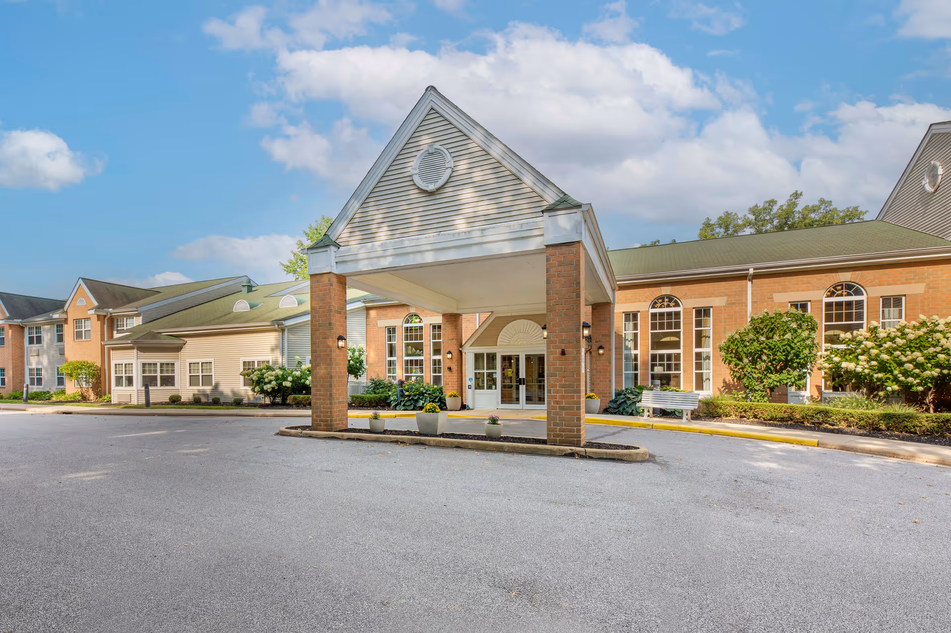 Exterior view of Brookdale Wickliffe senior living facility entrance with a covered drop-off area supported by brick columns, surrounded by well-maintained landscaping and a clear blue sky with some clouds.