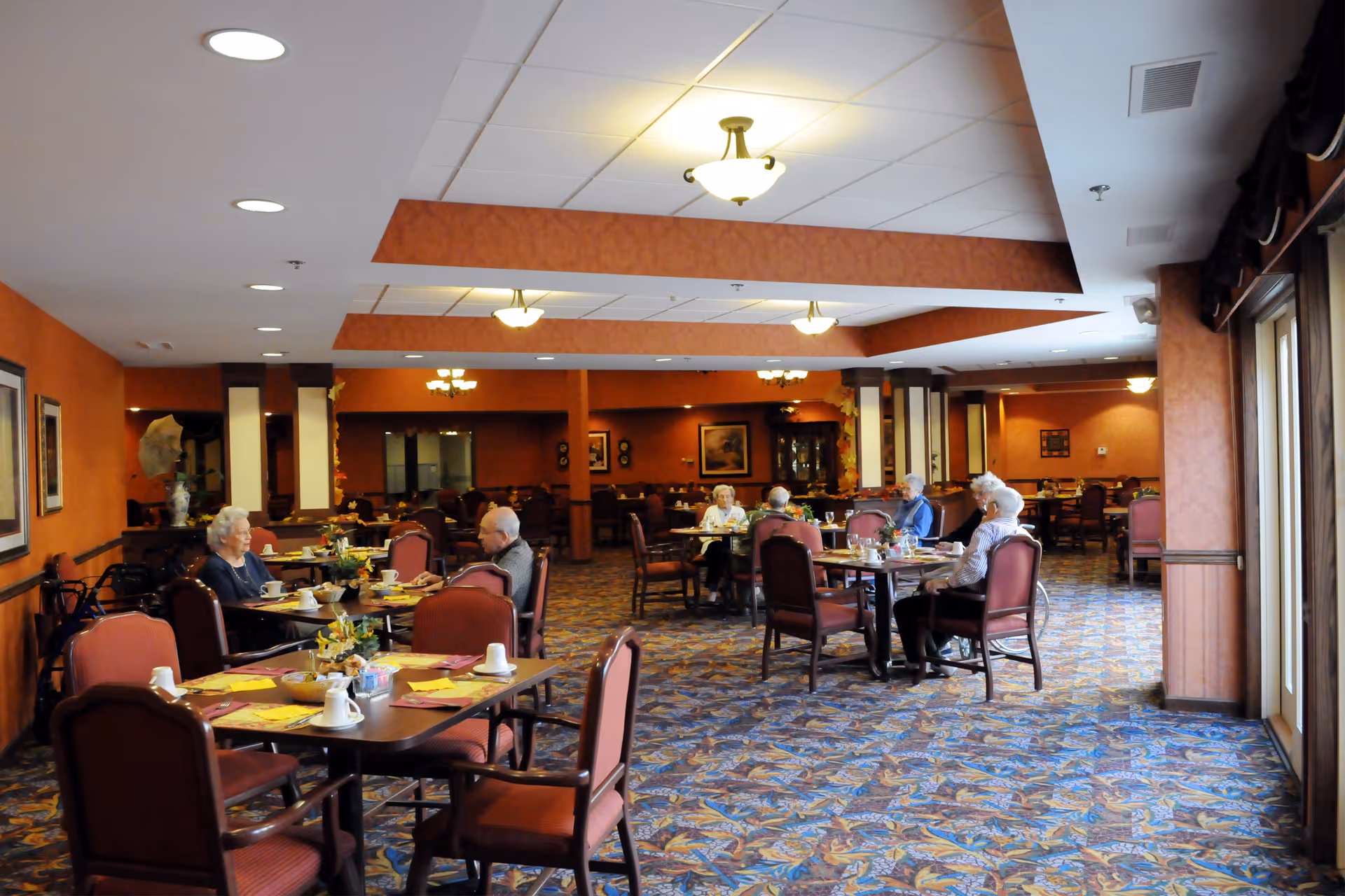 A dining room in a senior living facility with several elderly people seated at tables. The room has warm orange walls, patterned carpet, and ceiling lights. Tables are set with placemats, cups, and small flower arrangements.