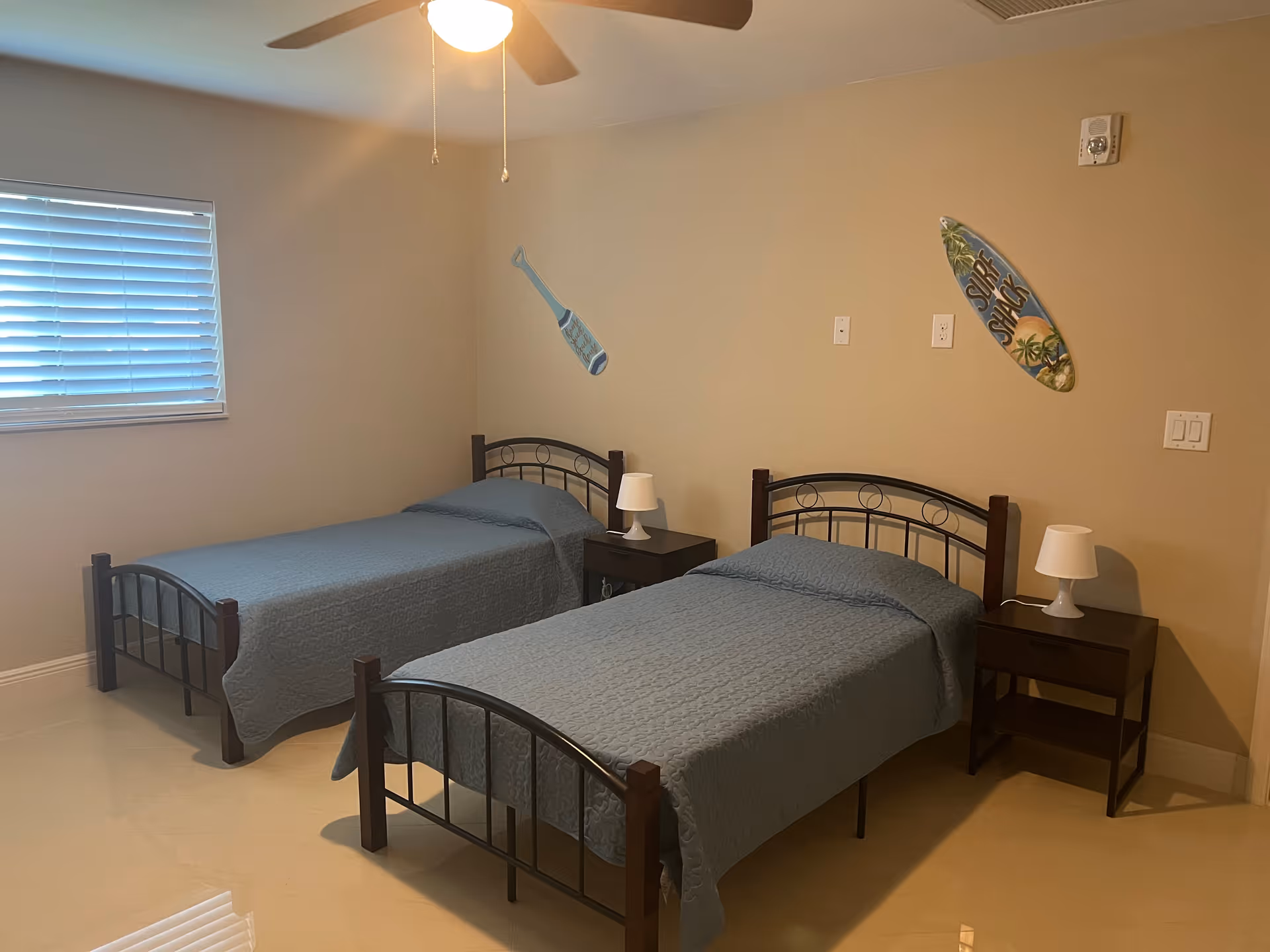 A bedroom with two twin beds covered in blue quilts, each with a dark wooden headboard and footboard. Between the beds is a dark wooden nightstand with a white lamp on it. The walls are beige with two decorative surfboard wall hangings. A window with white blinds is on the left wall, and a ceiling fan with a light is on the ceiling.