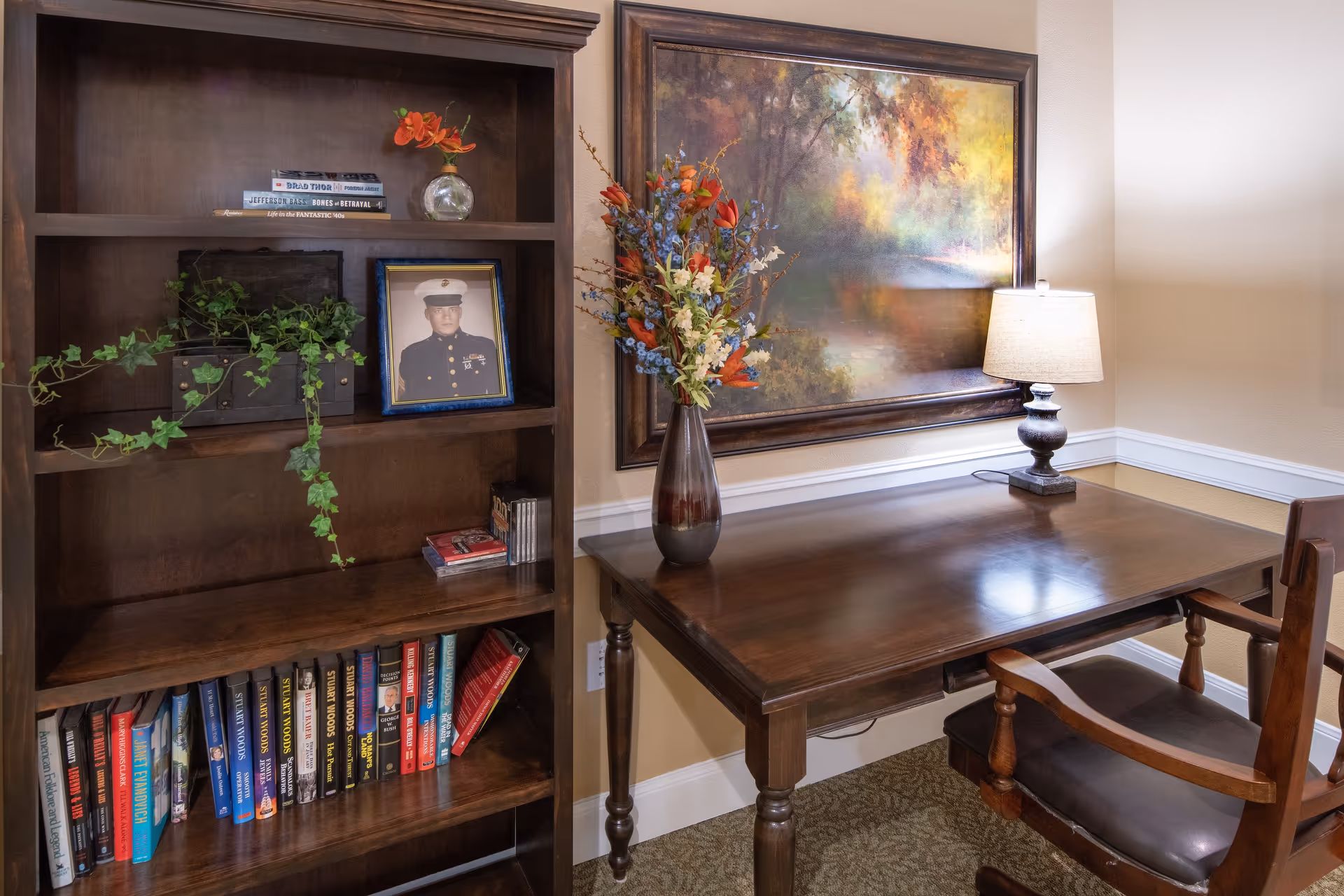 A cozy corner with a wooden desk and chair, a table lamp, a vase with colorful flowers, a large framed painting of a forest scene on the wall, and a wooden bookshelf filled with books, a framed photo of a man in military uniform, a small plant, and a glass vase with orange flowers.