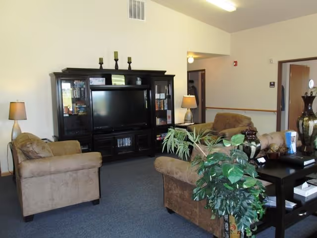 A cozy living room area with two brown upholstered armchairs and a matching loveseat arranged around a black entertainment center with a flat-screen TV. The entertainment center has glass cabinets with books and decorative items. There are two table lamps with beige shades on either side of the room, and a black coffee table with decorative vases and plants in the foreground. The walls are light-colored, and there is a doorway leading to another room.