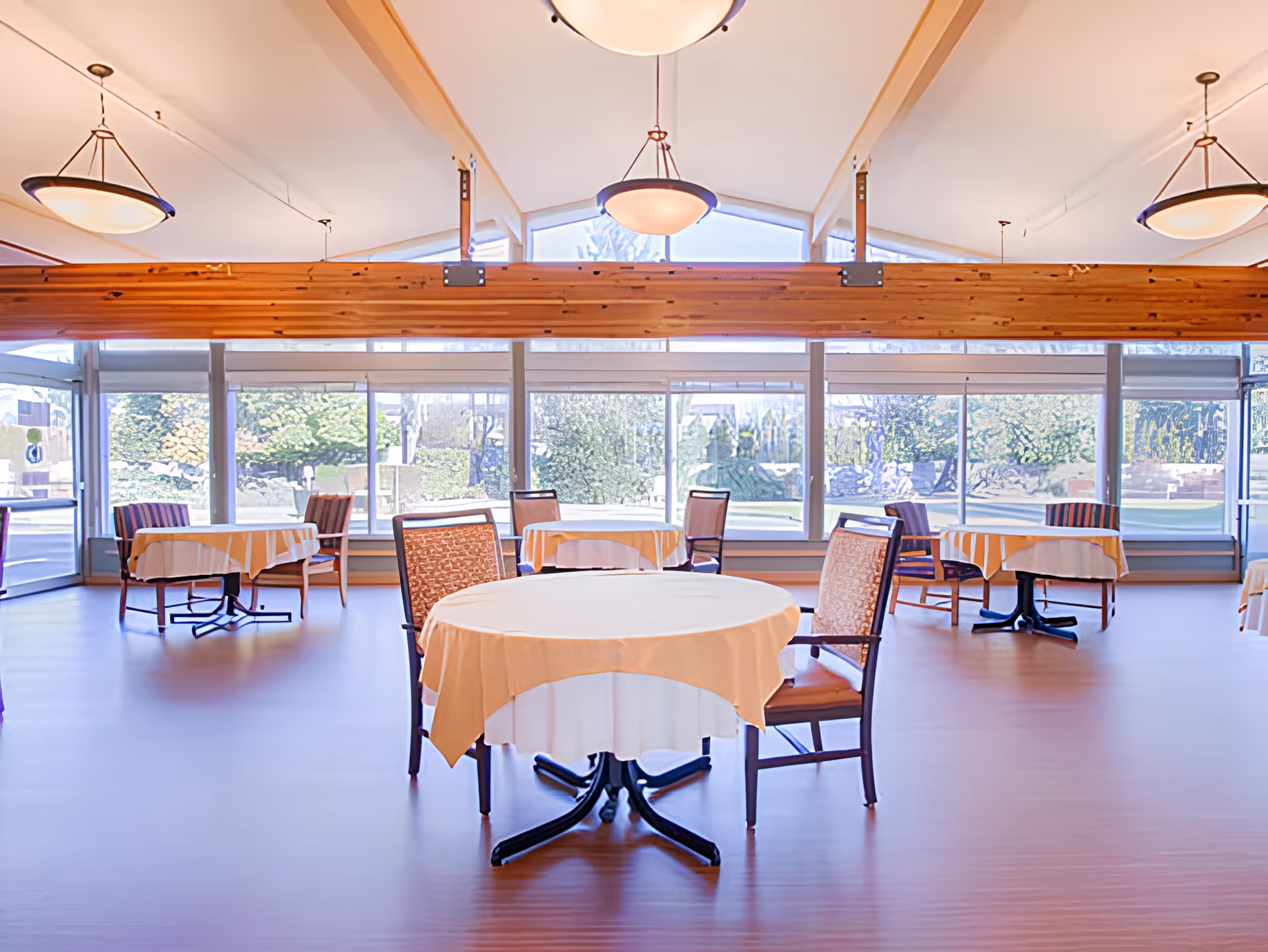 A bright dining room with round tables covered in white and yellow tablecloths, surrounded by wooden chairs with patterned cushions. Large windows line the back wall, allowing natural light to fill the space and offering a view of greenery outside. Ceiling lights hang from the high, vaulted ceiling with wooden beams.