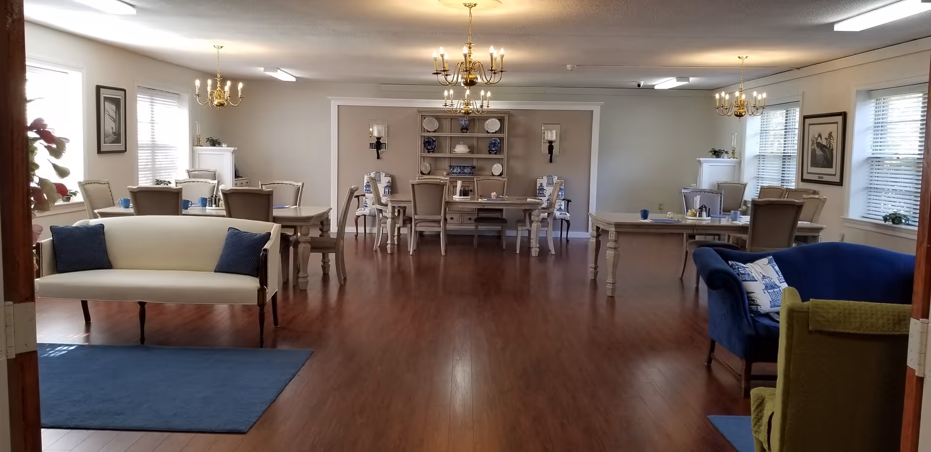 Spacious senior living dining and common room with multiple tables, chairs, sofas, chandeliers, and hardwood floors.
