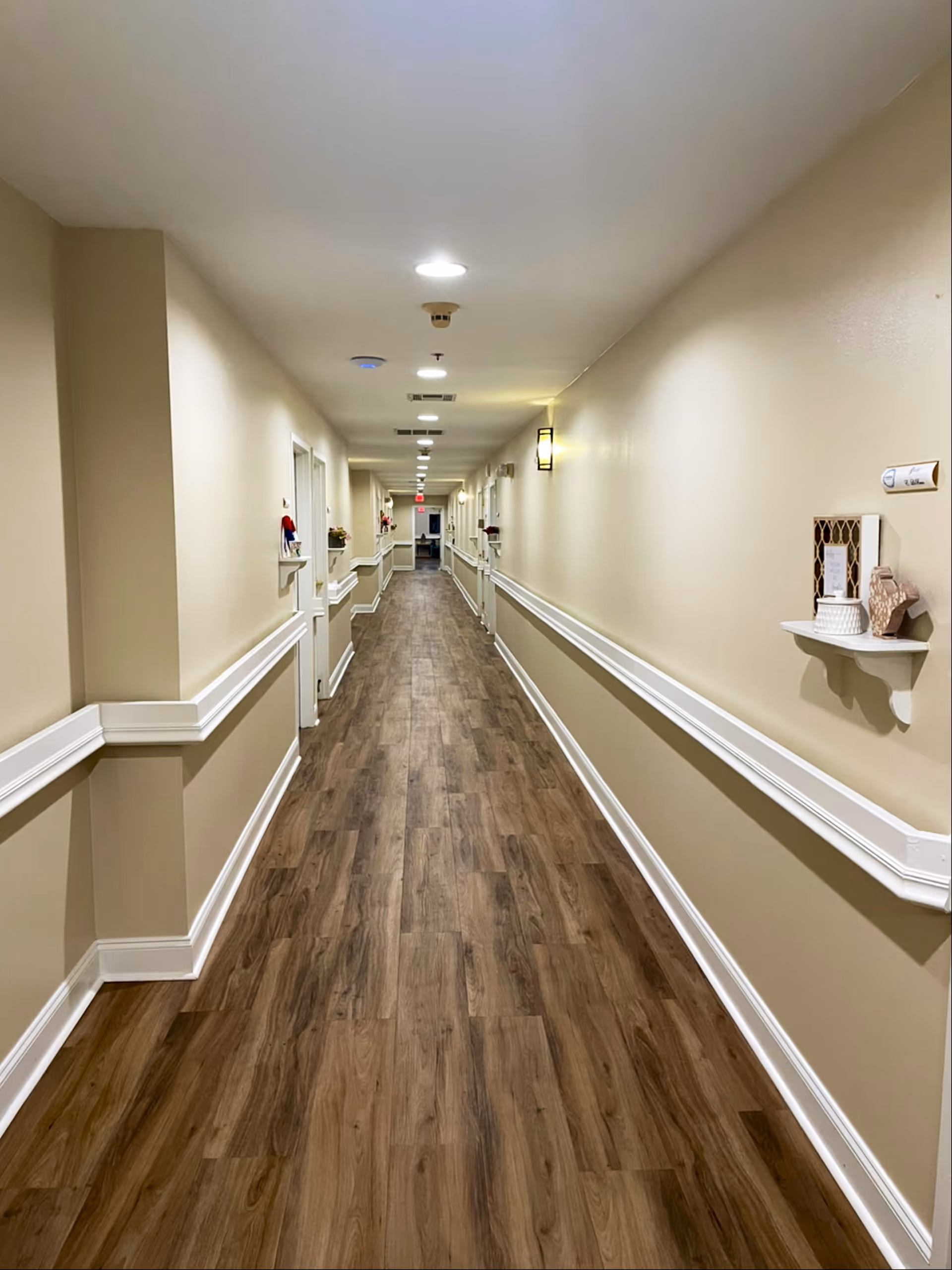 Long, well-lit hallway with wood-patterned flooring and beige walls featuring white trim and handrails. Small decorative shelves with items are mounted on the walls along the corridor, which has multiple doors on either side and ceiling lights evenly spaced.