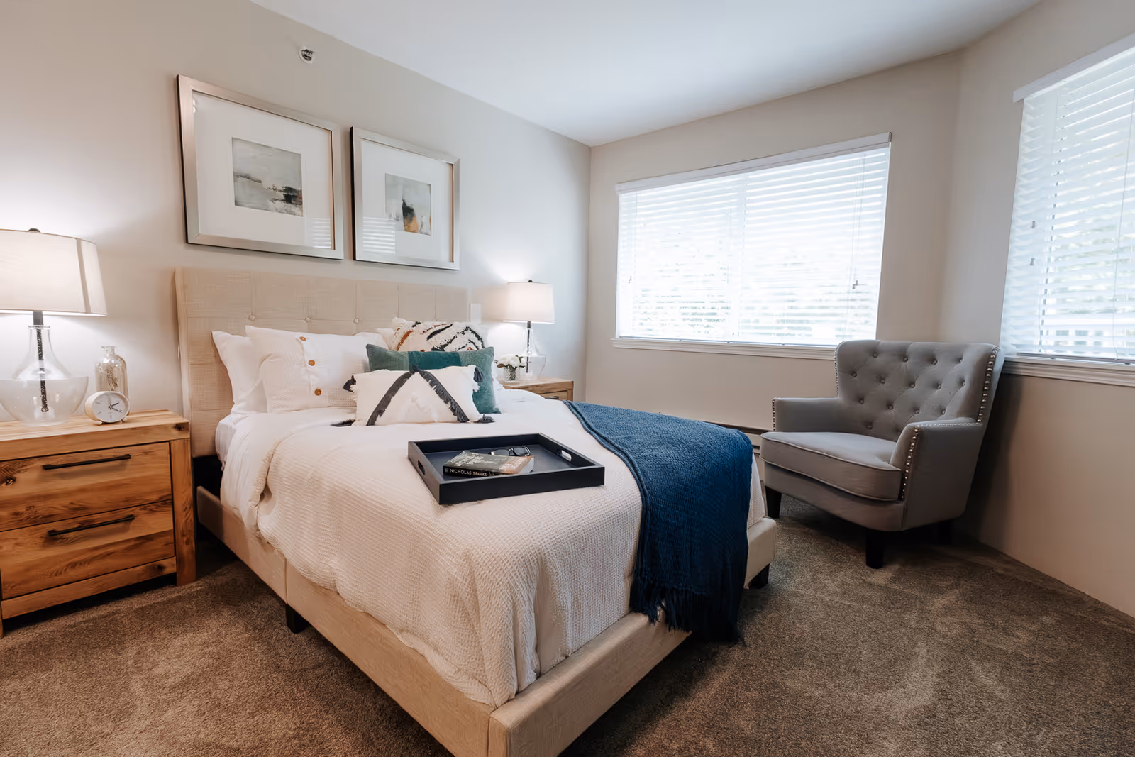 A cozy bedroom with a beige upholstered bed adorned with white and patterned pillows, a blue throw blanket, and a black tray on top. There are two wooden nightstands with lamps and decorative items on either side of the bed. A gray tufted armchair is positioned near two large windows with white blinds, and the room has beige walls and carpeted flooring.