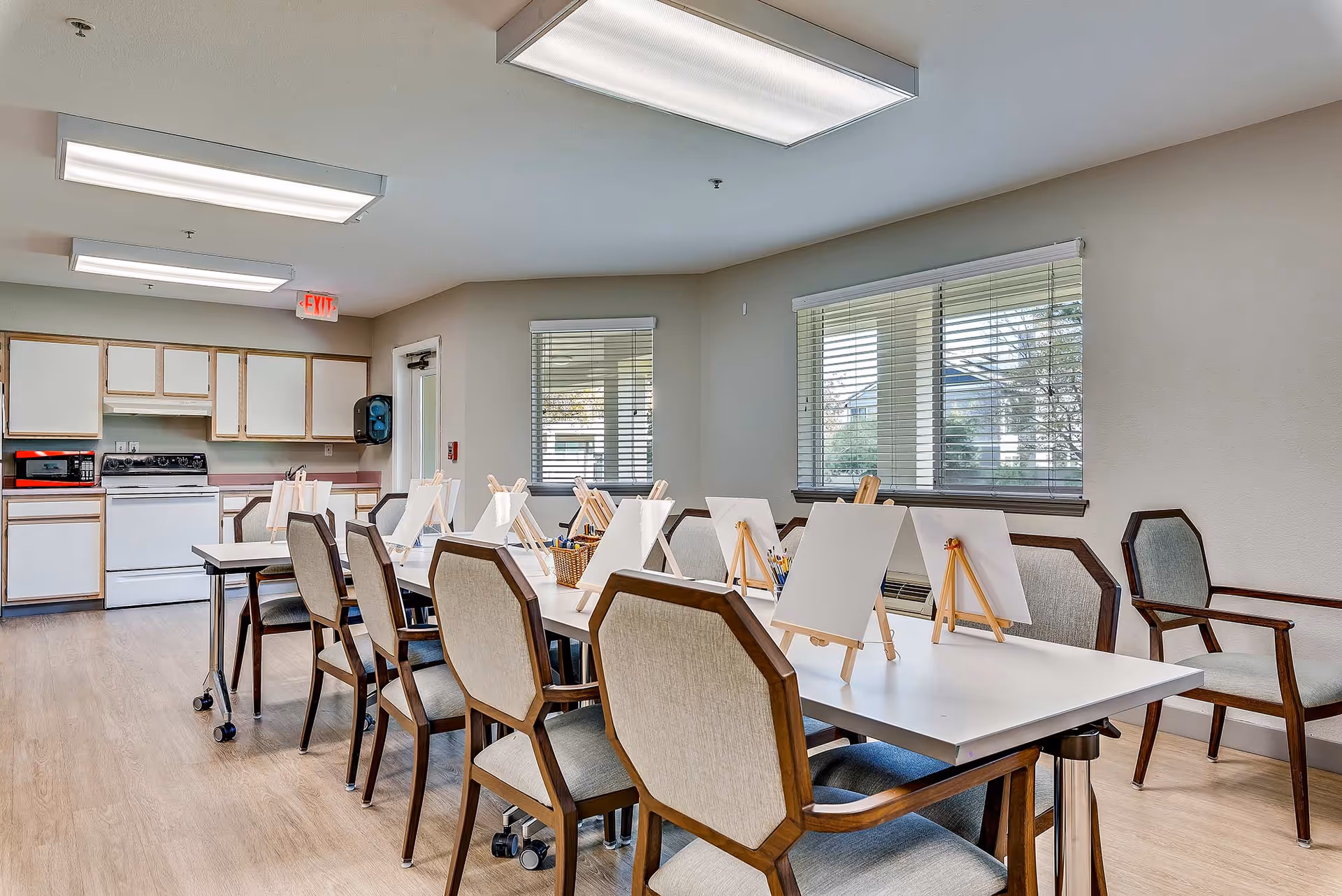 A bright activity room in a senior living facility with a long white table surrounded by cushioned wooden chairs. The table is set up with small easels holding blank canvases and baskets containing paintbrushes and art supplies. The room has large windows with blinds, light wood flooring, and a kitchenette area with white cabinets, a stove, and a microwave in the background.