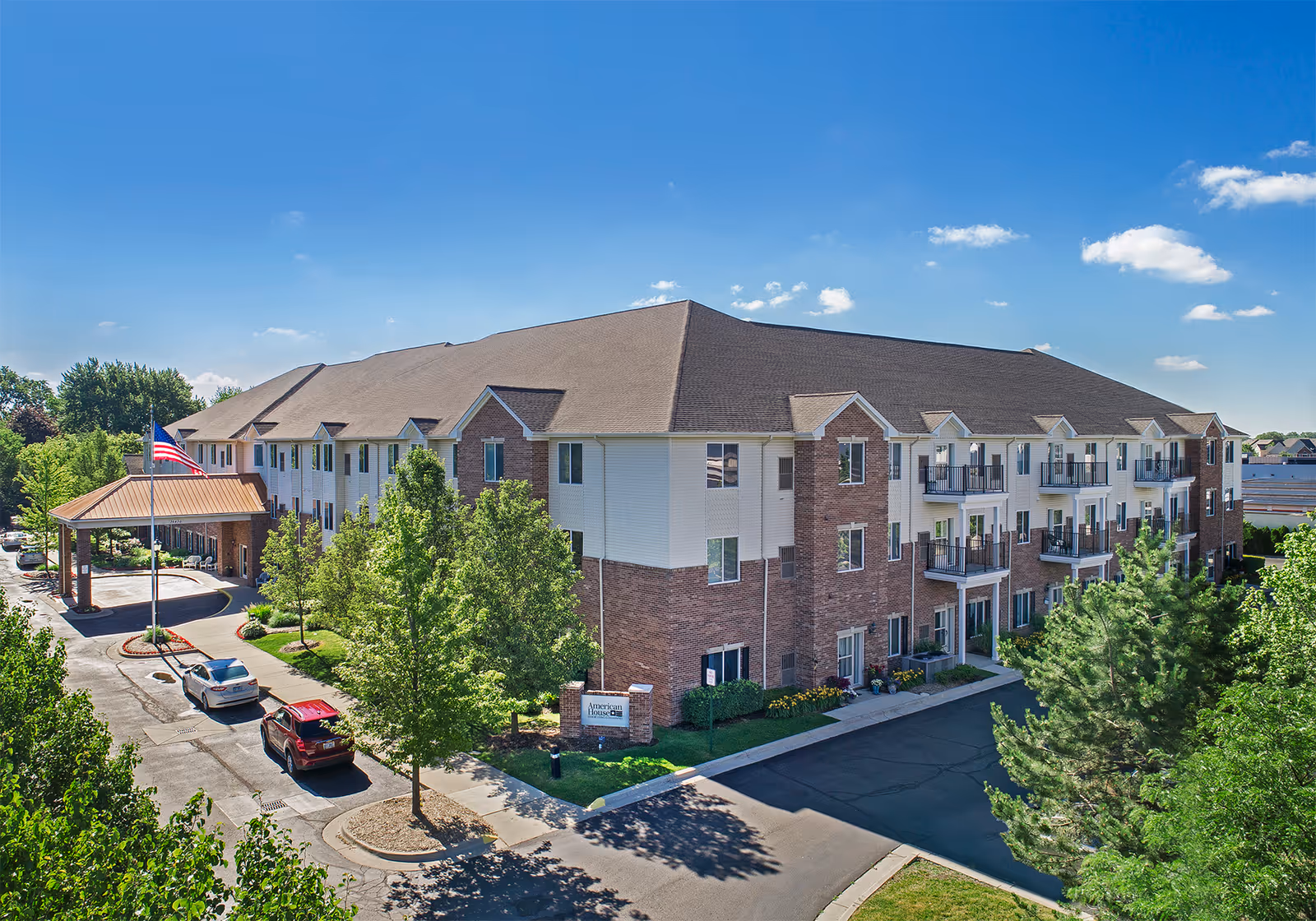Three-story brick and white-siding senior living building with balconies, a covered entrance and American flag, surrounded by trees and parked cars.