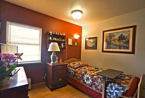 Bedroom in ARIA of Waukesha with single bed, quilt, nightstand, lamp, shelf, and framed art.
