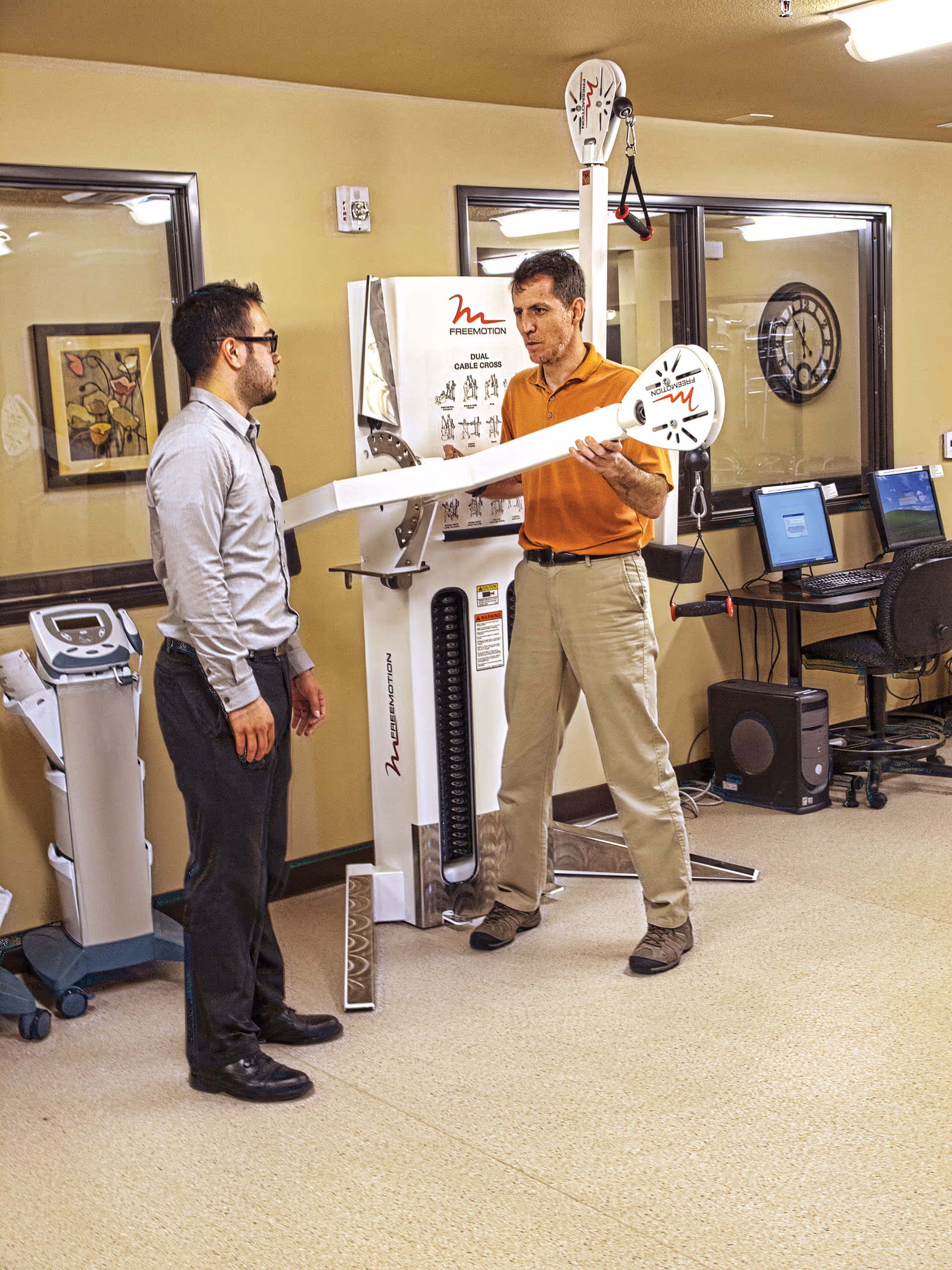 Two men in a rehabilitation or physical therapy room. One man in an orange shirt is demonstrating or adjusting a piece of exercise equipment labeled 'FREEMOTION' to another man wearing glasses and a light gray shirt. The room has beige walls, framed artwork, a clock, and computer workstations in the background.