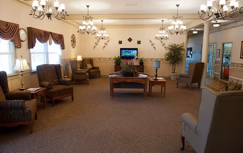 A spacious living room area with multiple upholstered armchairs arranged along the walls and around a central wooden coffee table with plants and magazines. The room features beige walls with decorative trim, several chandeliers hanging from the ceiling, large windows with brown valances, and a flat-screen TV mounted on the far wall. There is a large mirror on the right wall and a potted plant near the TV.
