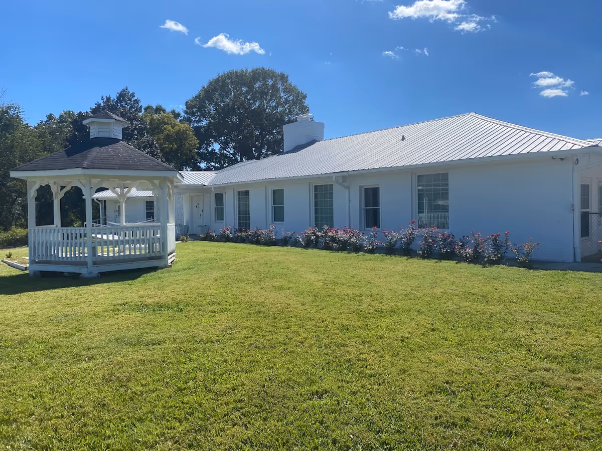 White single-story building with a metal roof and a white gazebo on a grassy lawn under a blue sky.