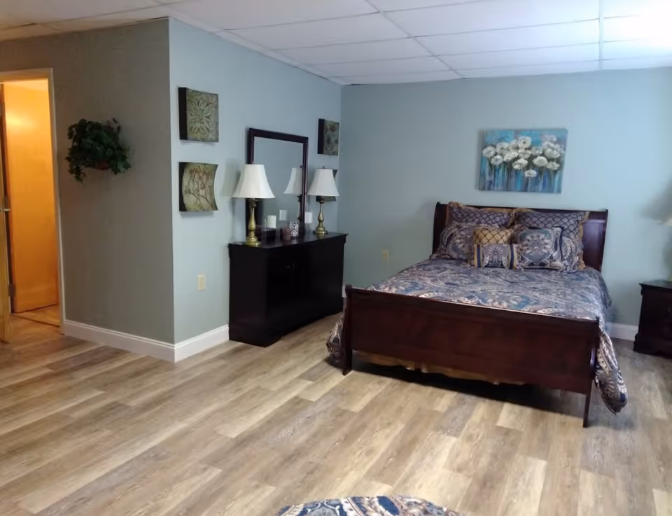 A bedroom with a wooden bed frame and a bed made with patterned blue and beige bedding. There is a matching wooden dresser with two lamps and a mirror above it. The walls are painted light blue and decorated with floral artwork. The floor is wood laminate, and there is an open doorway leading to another room.