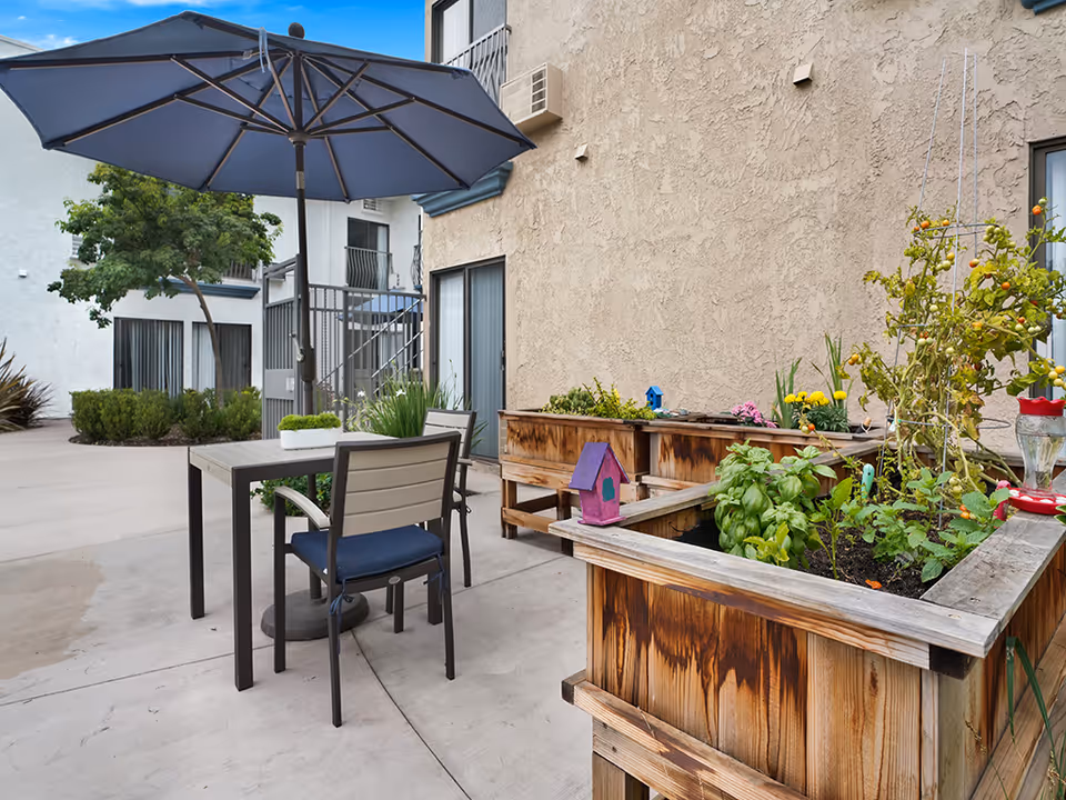 Outdoor patio area with a table and two chairs under a large blue umbrella. There are raised wooden garden beds with various plants and flowers growing. The patio is adjacent to a beige stucco building with sliding glass doors and windows.