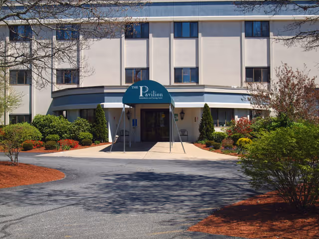 Front exterior view of The Pavilion Rehabilitation and Nursing Center building with a green awning over the entrance, surrounded by landscaped bushes and trees.