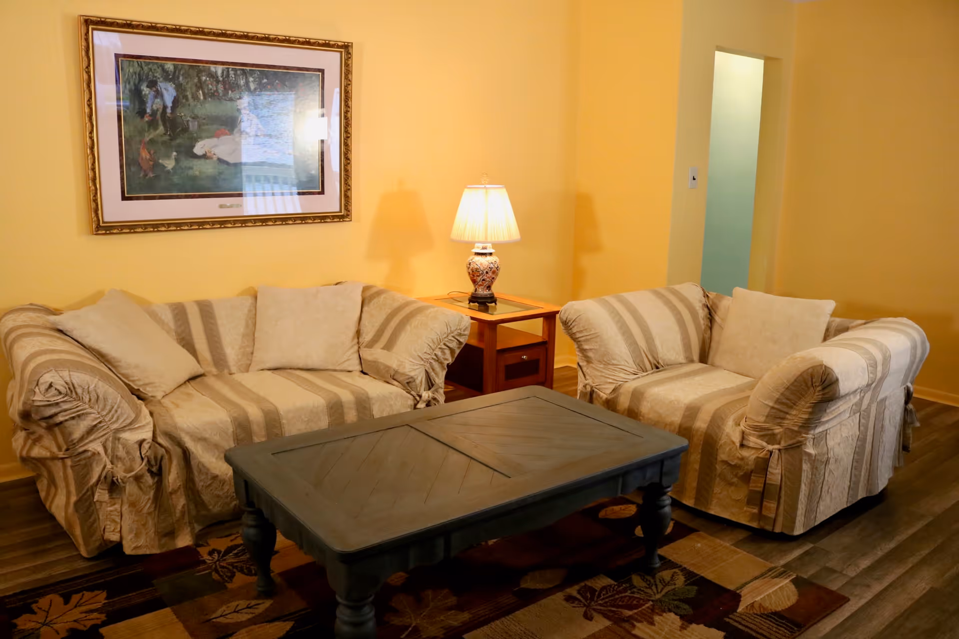 A cozy living room with two beige striped sofas, a wooden coffee table, a side table with a decorative lamp, and a framed painting on a yellow wall.