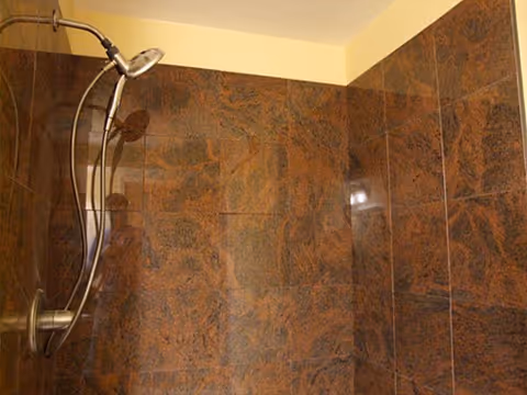 Shower area with dark brown and black marbled tiles on the walls and a silver handheld showerhead attached to a curved metal rod.