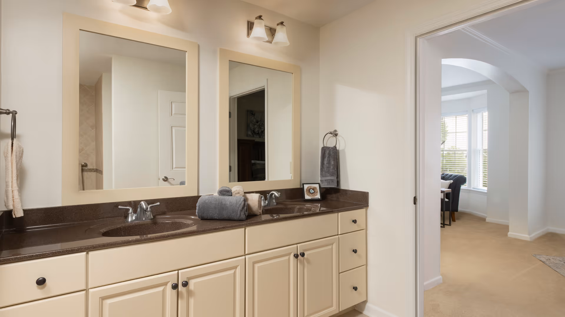 Double-sink bathroom vanity with brown countertop, two mirrors, towels, and a doorway opening to a carpeted room.