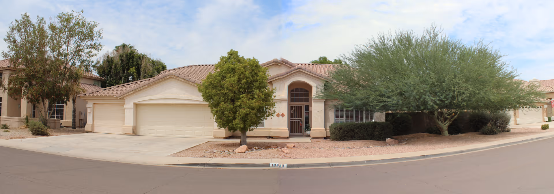 Single-story residential building with a beige exterior, tiled roof, and a two-car garage. The front yard features desert landscaping with rocks, a small tree, and a larger tree near the entrance. The sky is partly cloudy.