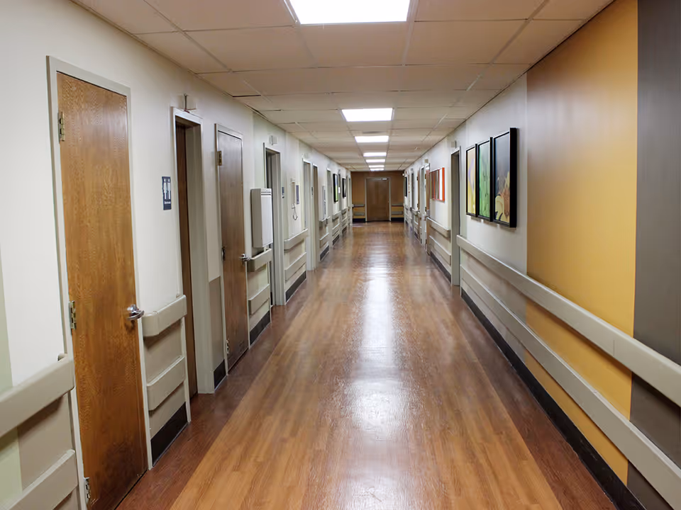 A long, clean hallway in a senior living facility with wooden doors on both sides, handrails along the walls, framed artwork on the right wall, and bright overhead lighting.