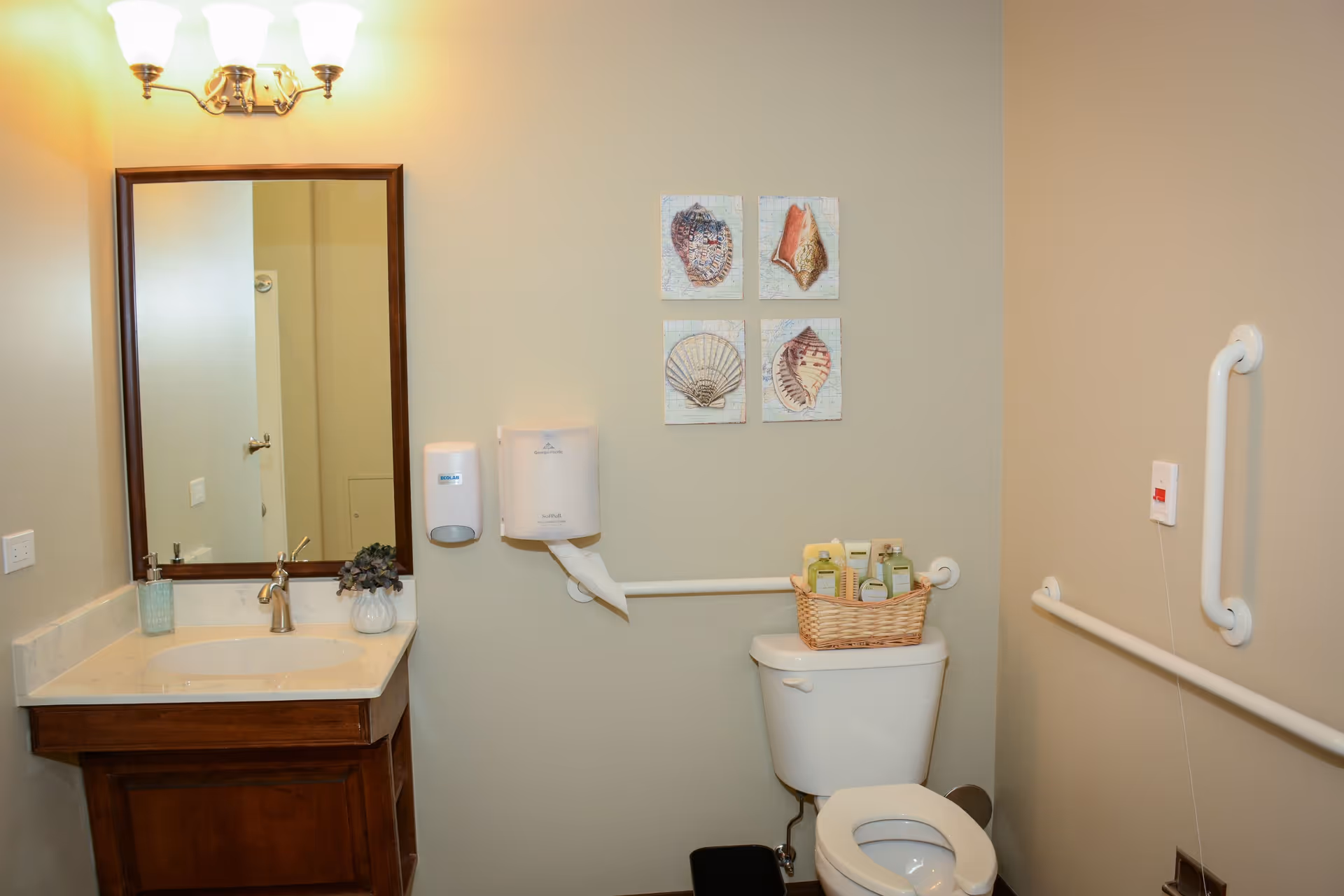 A clean bathroom with beige walls featuring a wooden vanity with a white sink and a mirror above it. There is a wall-mounted soap dispenser and paper towel holder next to the sink. Above the toilet, there is a basket with toiletries and four framed pictures of seashells on the wall. Grab bars are installed on the walls near the toilet for accessibility.