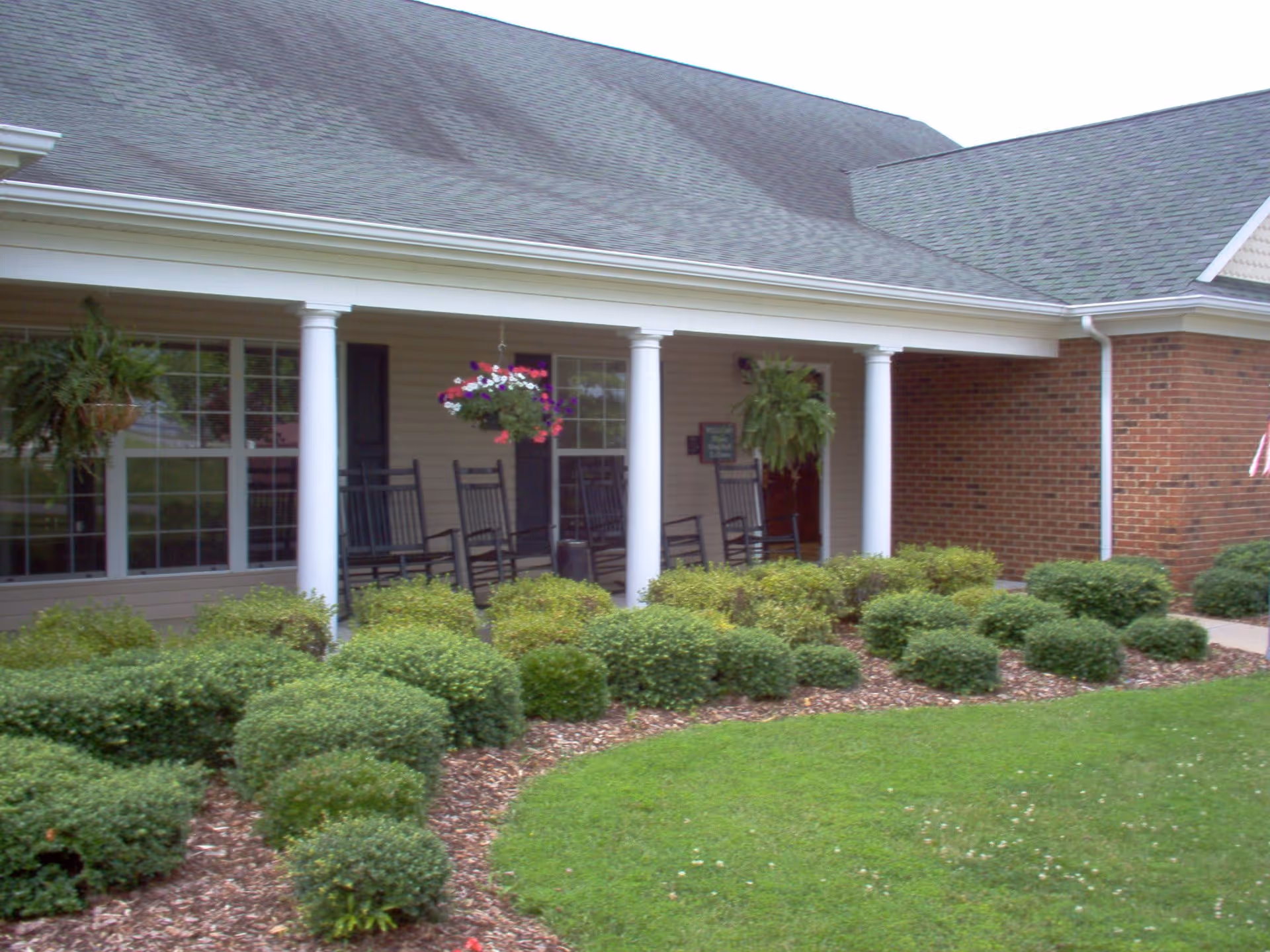 Front porch of a building with white columns, hanging flower baskets, black rocking chairs, and neatly trimmed bushes in the garden bed in front of the porch.