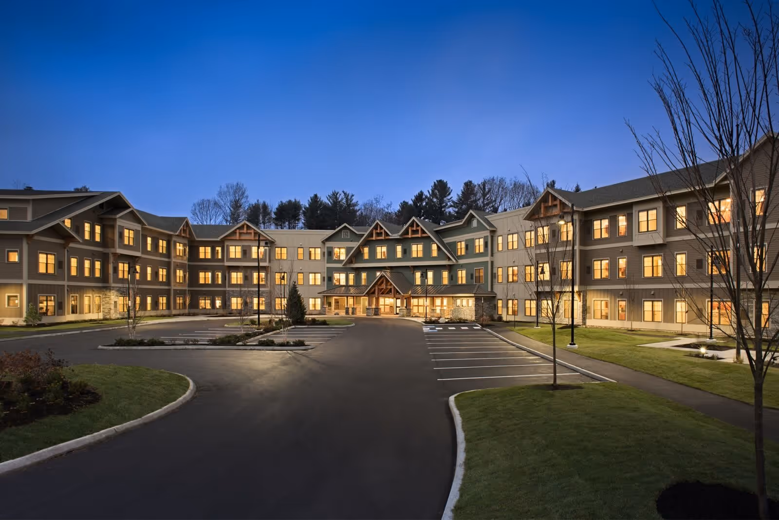 Exterior view of Stroudwater Lodge at dusk showing a large, three-story building with multiple lit windows, a paved driveway, parking spaces, and landscaped grassy areas with young trees.
