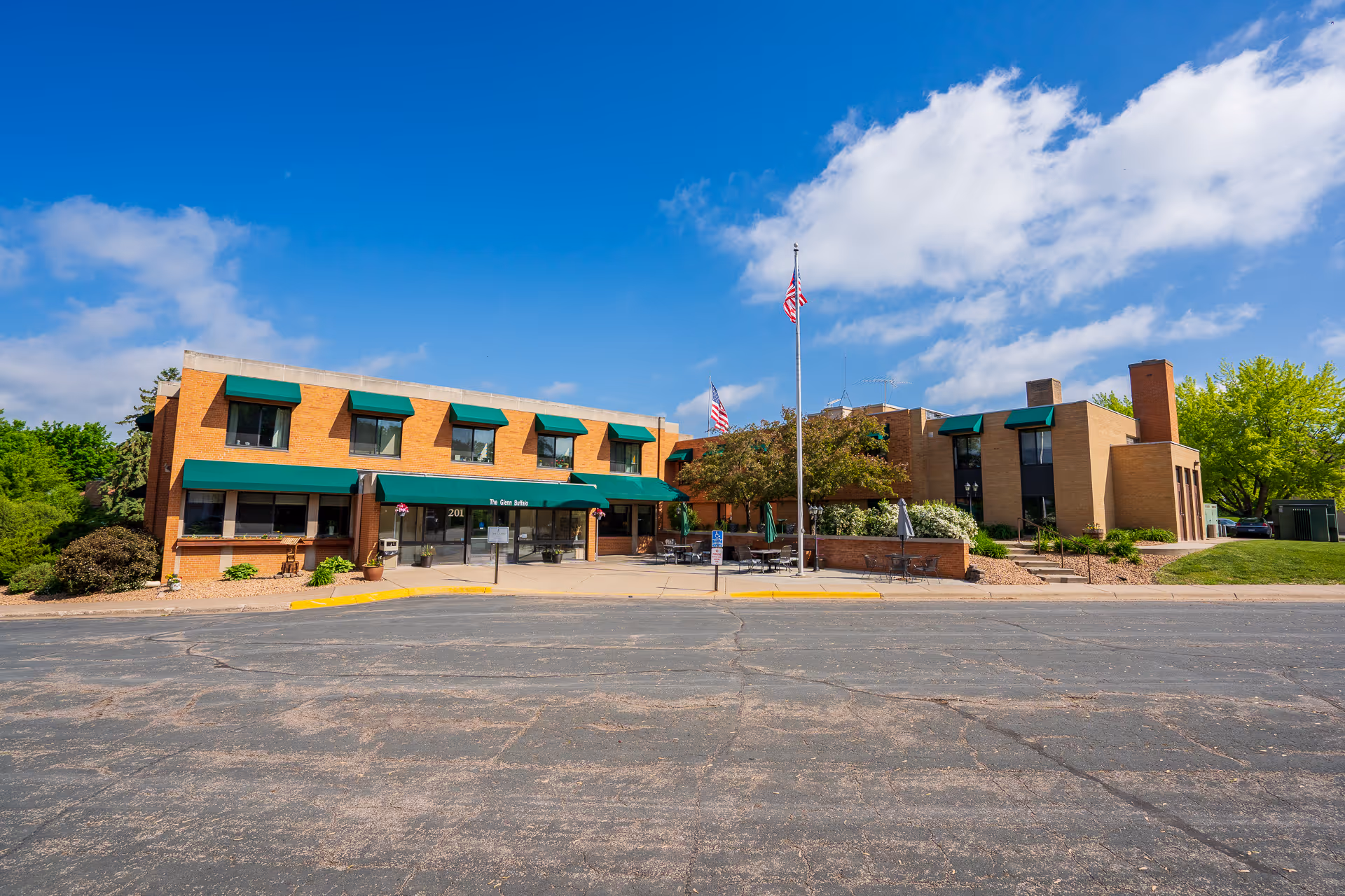 Exterior view of The Glenn Buffalo senior living facility building under a blue sky with some clouds. The building is two stories with a brick facade and green awnings over the windows and entrance. There are two American flags on flagpoles in front of the building, along with some outdoor seating and landscaping.
