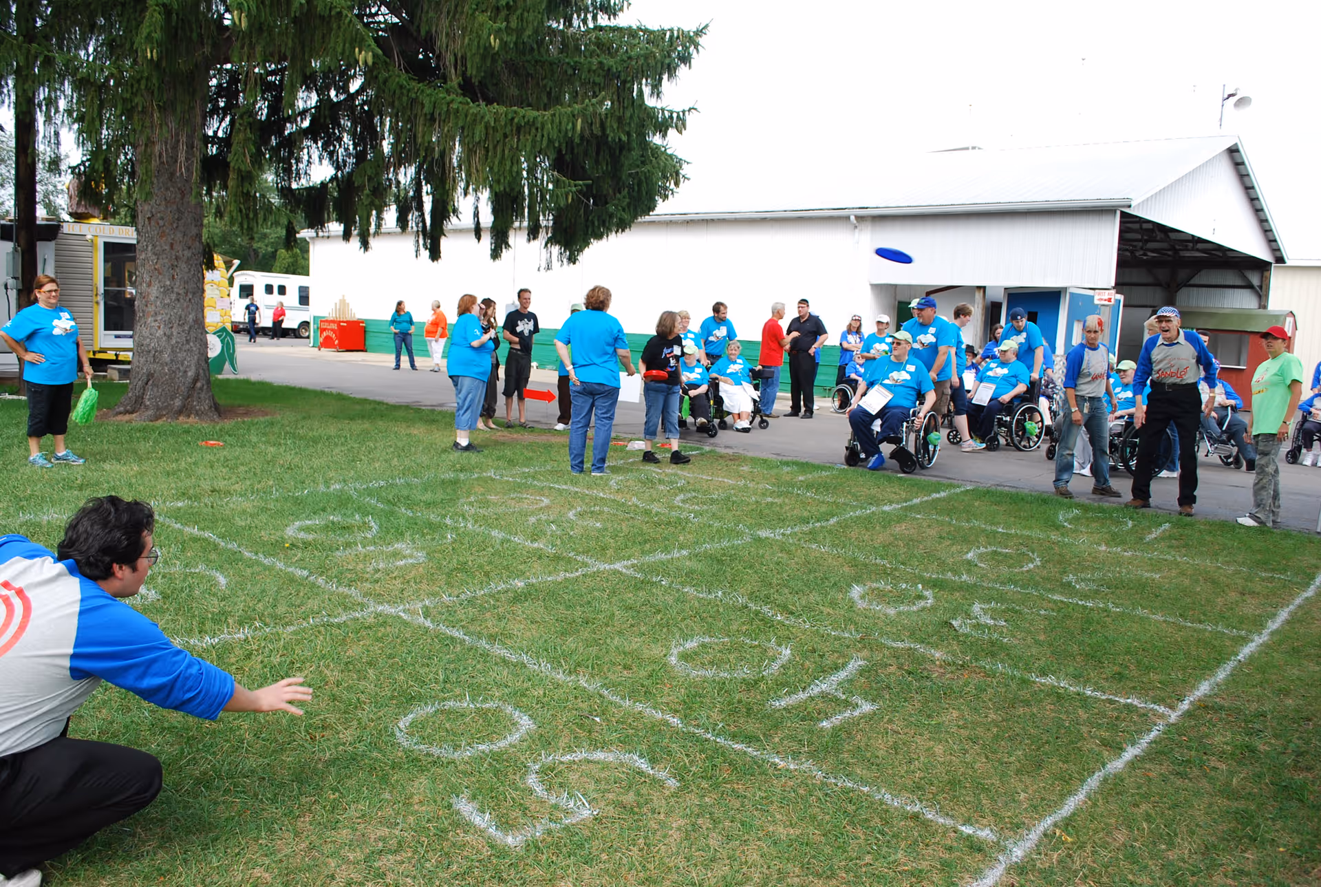 A group of people, some in wheelchairs and others standing, gathered outdoors on a grassy area with a large tree. They are playing a lawn game with a grid marked on the grass and a blue frisbee in mid-air. Many participants are wearing matching blue shirts. There are buildings and a few more people in the background.