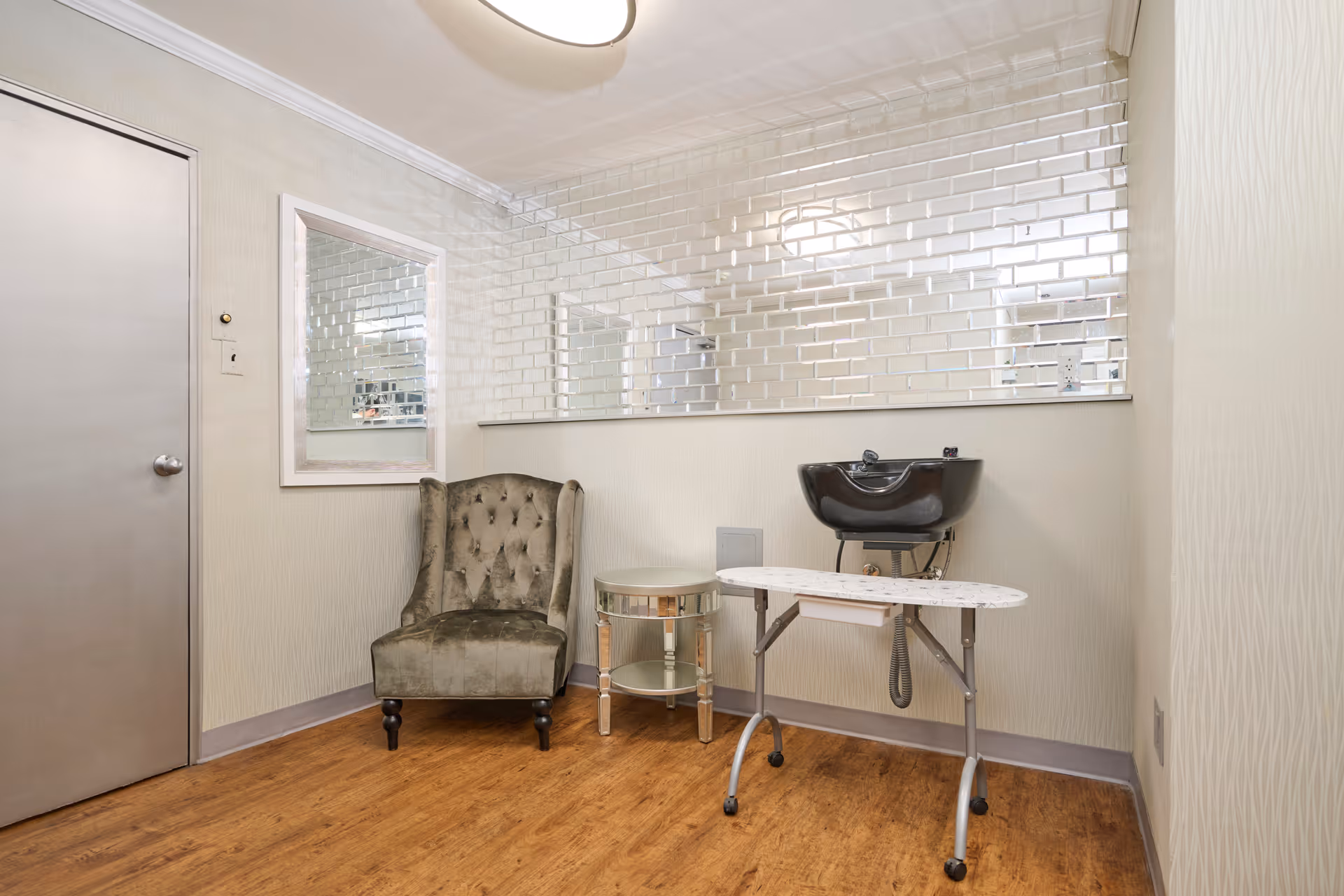 Interior salon corner with a tufted armchair, mirrored tile wall, black shampoo sink and a small rolling styling table on wood floors.
