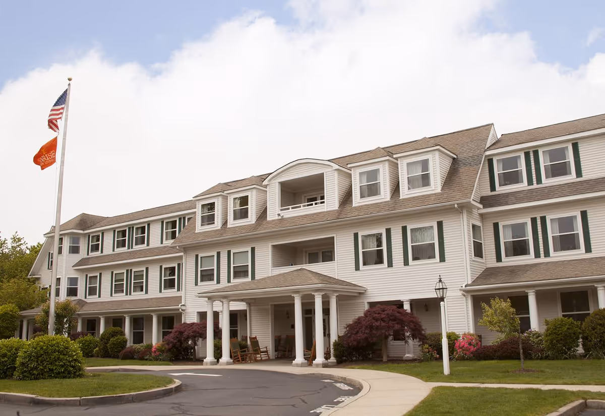Exterior view of a large, white three-story senior living facility building with multiple windows and green shutters. The entrance has a covered porch with white columns and rocking chairs. There are well-maintained bushes, small trees, and a curved driveway in front. Two flags, including an American flag, are flying on a flagpole to the left.