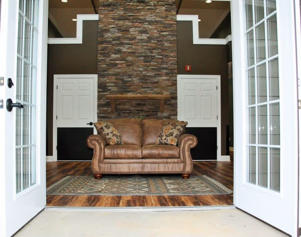 View through open double glass doors into a cozy interior space featuring a brown leather loveseat with patterned cushions, placed on a decorative rug in front of a tall stone fireplace with a wooden mantel. The room has dark brown walls, white trim, and two white doors on either side of the fireplace.