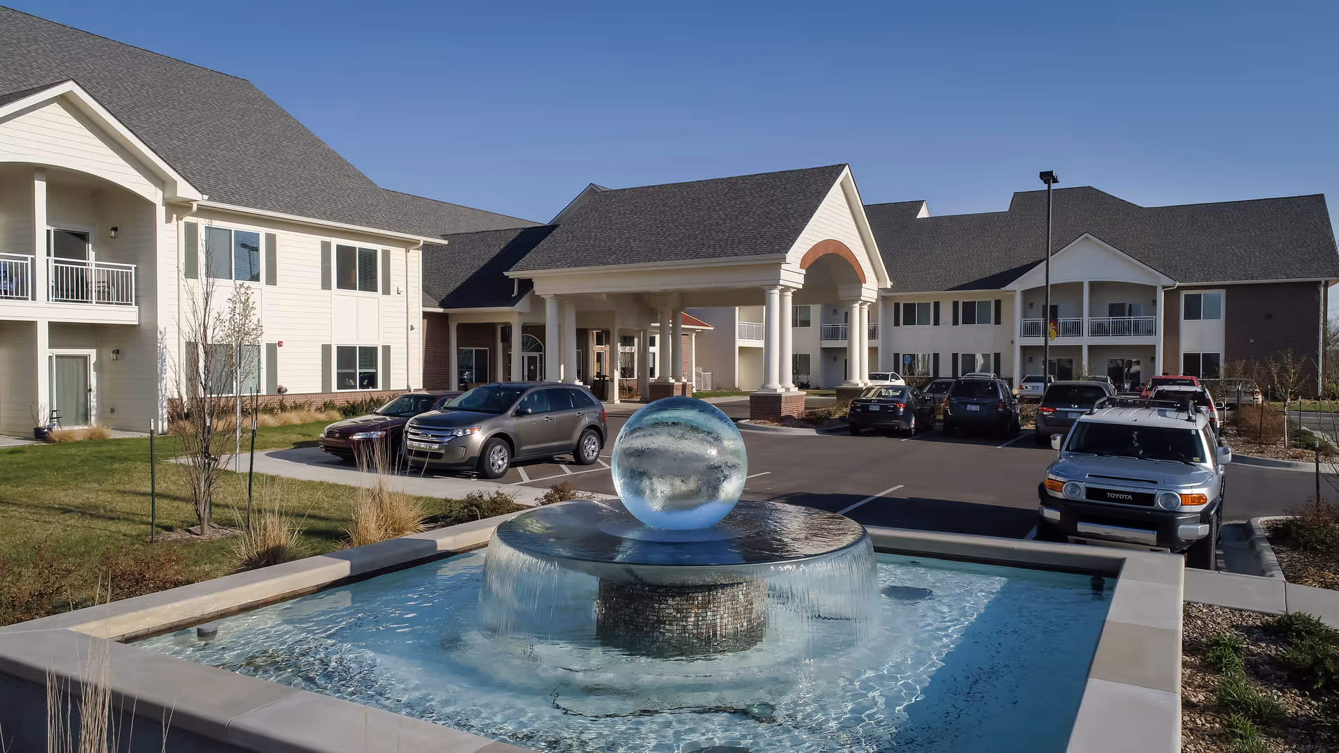 Exterior view of Pioneer Ridge Independent Living of Lawrence showing a large water fountain with a glass sphere in the center, surrounded by a pool of water. Behind the fountain is the entrance to the building with a covered portico supported by columns. Several cars are parked in the parking lot in front of the building, and the sky is clear and blue.